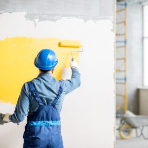 Workman in uniform painting wall with yellow paint at the construction site indoors